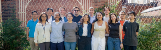 14 members of bewersdorf lab stand in two lines in front of greenery and white gate, smiling at the camera