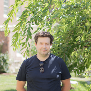 headshot of florian schueder, caucasian man with short brown hair in navy t shirt with hands behind back looking at the camera