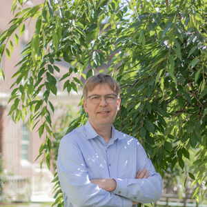 caucasian man with shirt blond hair and glasses in light blue button up shirt with arms crossed across chest smiling at camera standing outside in front of a tree