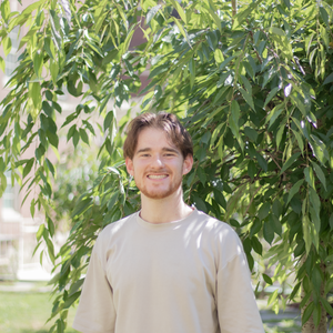 caucasian man with short reddish hair wearing a tan t shirt with hands in pockets smiling at the camera standing outside in front of a tree