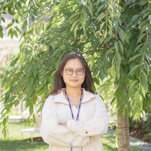 east asian woman with long brown hair and black glasses wearing a beige jacket with arms crossed across chest smiling at the camera standing outside in front of a tree