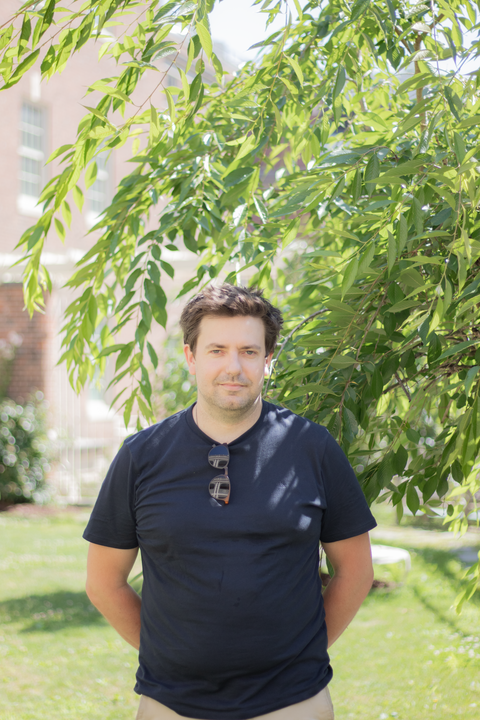 headshot of florian schueder, caucasian man with short brown hair in navy t shirt with hands behind back looking at the camera