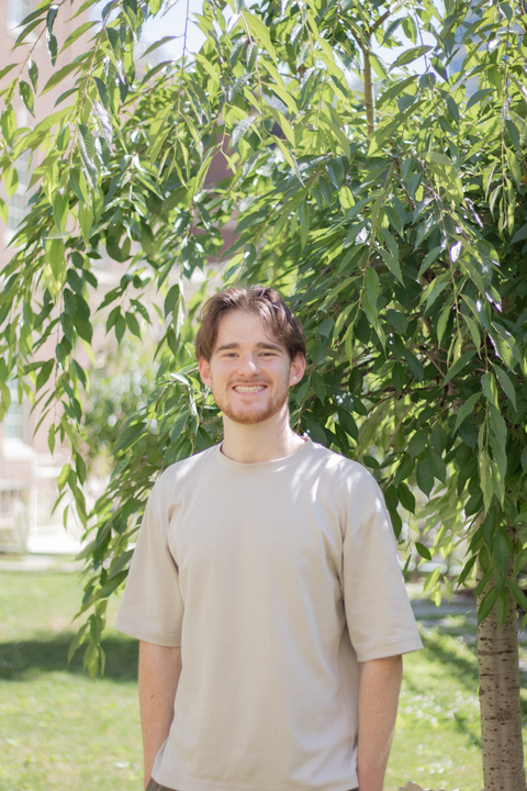 caucasian man with short reddish hair wearing a tan t shirt with hands in pockets smiling at the camera standing outside in front of a tree