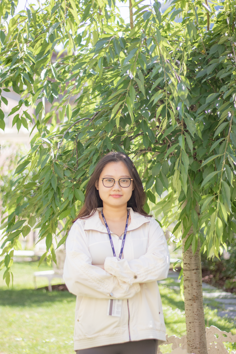 east asian woman with long brown hair and black glasses wearing a beige jacket with arms crossed across chest smiling at the camera standing outside in front of a tree